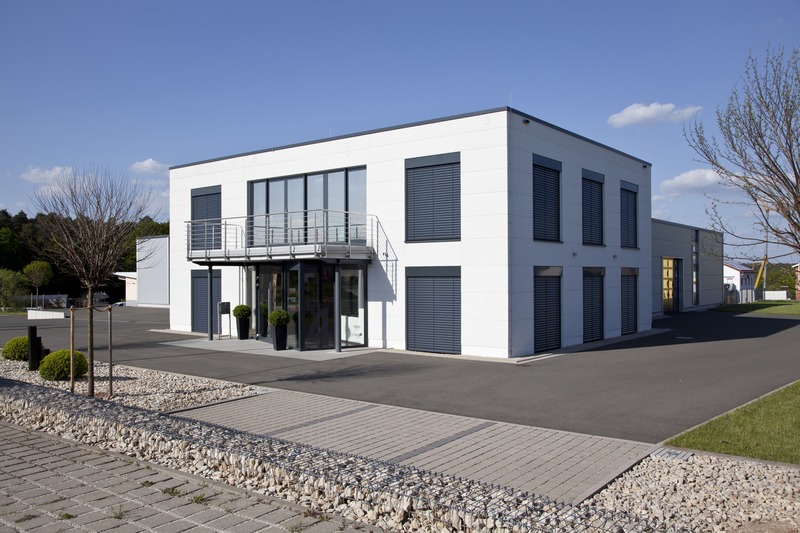 Modernes zweigeschossiges Bürogebäude mit weißer Fassade, anthrazitfarbenen Raffstore-Verschattungen, Balkon mit Edelstahlgeländer und gepflastertem Vorplatz vor blauem Himmel